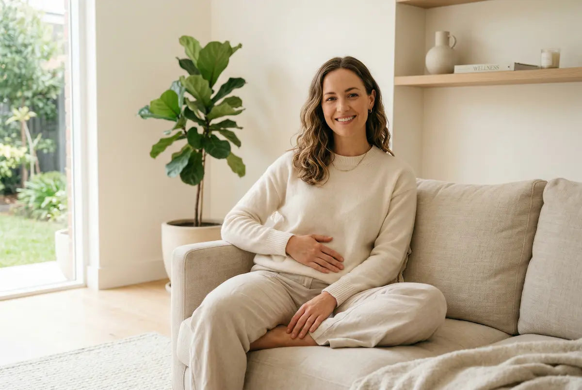 Smiling woman sitting comfortably on a sofa at home, hand resting on her abdomen, conveying relief from digestive discomfort.