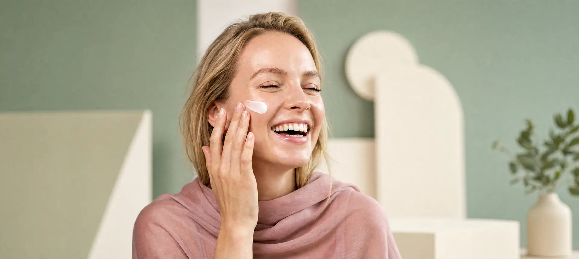 A smiling woman with freckles applying a dab of white face cream to her cheek. Clean skin, natural aesthetic, and bright lighting