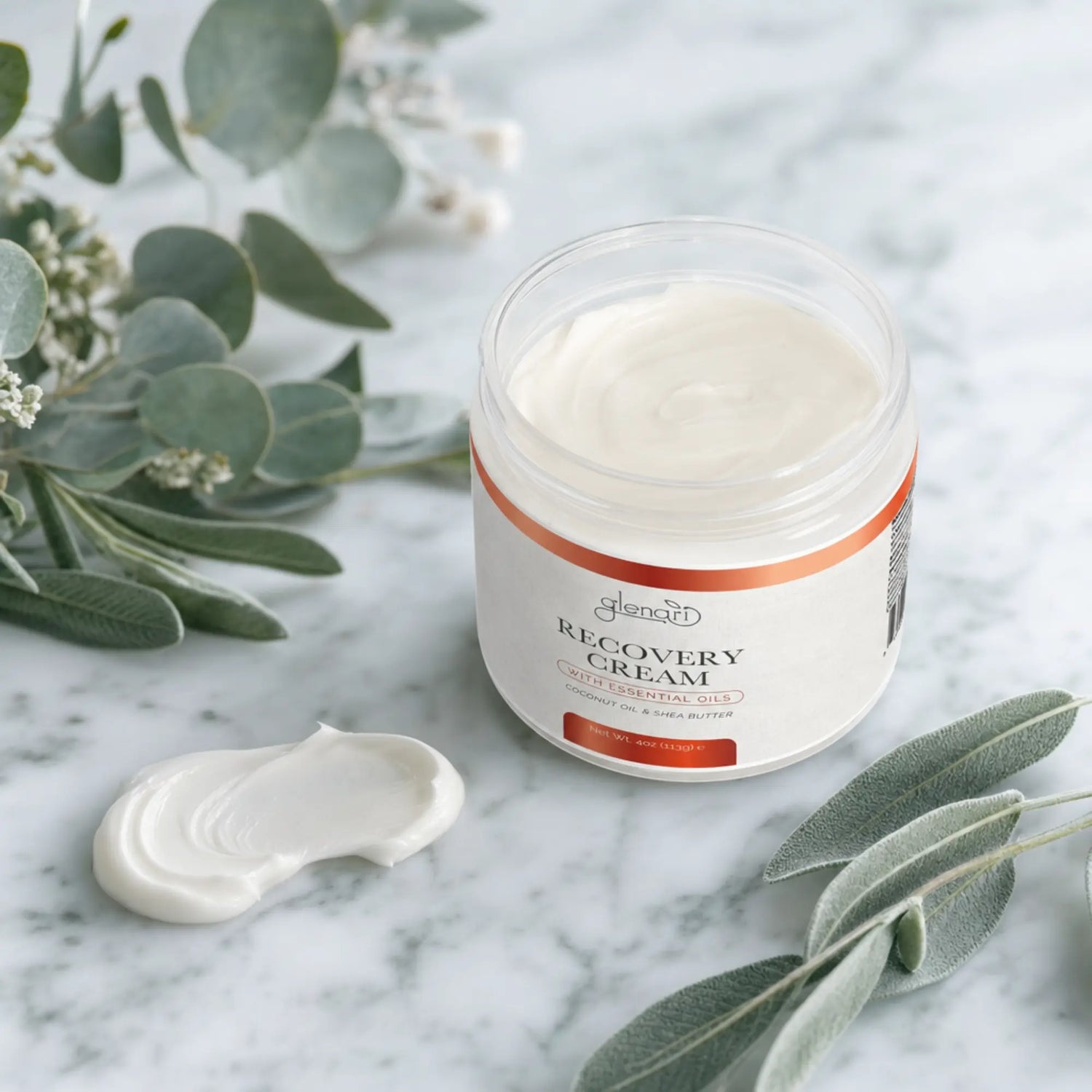 An open jar of Glenari Recovery Cream sits on a white marble surface with a dollop of the white cream beside it and soft green botanical leaves around it.